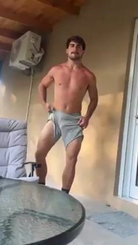 A shirtless man wearing gray shorts and black socks flexes his muscles outside on a patio. An air conditioning unit is affixed to the wall behind him. To the left of the man, there is a gray patio chair and a glass-topped table. The patio is covered by a wooden roof. There is a white doorframe to the right of the man.