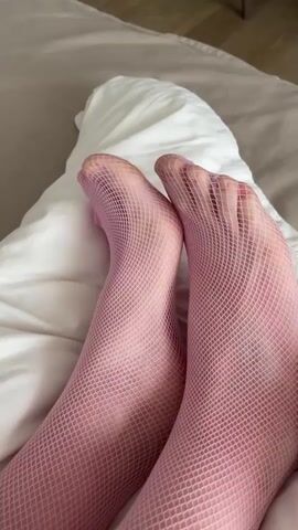 The video shows a close-up view of a person's feet, which are covered in pink fishnet stockings. The feet are resting on a white pillow, which is on a bed. The camera angle is from above, looking down at the feet. The lighting is soft and natural. The video is short, lasting only a few seconds, and there is no audio.