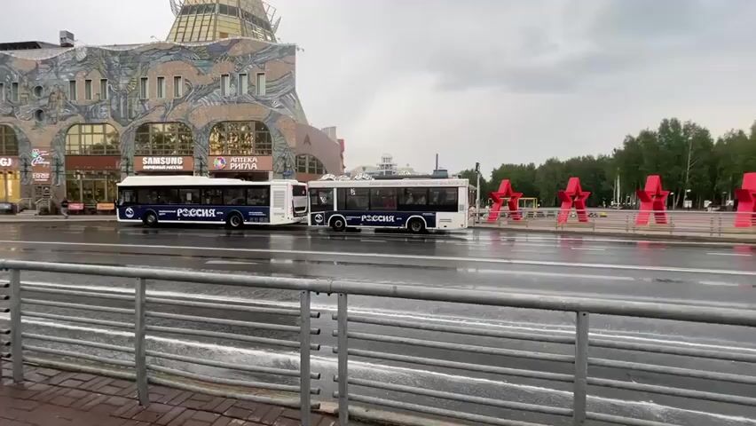 A video shows a cloudy day with two buses parked on a wet street in front of a building with a distinctive mosaic facade and a golden, conical roof. The buses are white with dark windows and have "РОССИЯ" (Russia) written on their sides in blue letters. A metal railing runs along the bottom of the frame, separating a sidewalk from the street. In the background, red star-shaped sculptures are visible.