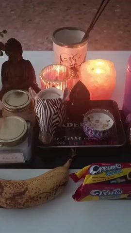 A table holds a collection of objects.  There are various candles, some lit, as well as incense sticks in a ceramic container. A small zebra head figurine sits next to a black crystal on a small tray with text on it. A banana with brown spots and a package of "Cream-O" cookies are positioned in the lower foreground. Also on the table are a statue of Buddha and several other decorative items. The scene is dimly lit. The table sits in front of a dark colored cabinet or wall.  The floor is a beige speckled surface. A small potted plant sits in the top left corner.