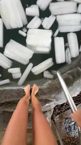 A woman in a black swimsuit enters a small pool filled with ice chunks. The pool is surrounded by stone and vegetation, and is under a thatched roof. The woman expresses shock as she gets in the pool.