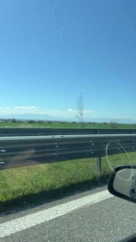 The video shows a view from a car moving along a highway on a clear, sunny day. The camera is positioned on the passenger side, looking out at the landscape. A gray metal guardrail runs alongside the highway. Beyond the guardrail is a field of green grass with a few bare trees and some distant hills or mountains on the horizon. The sky is bright blue with a couple of faint, circular smudges on the window visible in the frame. A side mirror of the car is visible in the lower right corner of the frame. The asphalt road is gray with white lane markings.