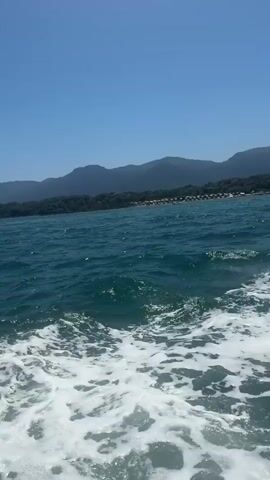The video shows a view from a boat moving through blue water on a clear day. The camera is angled downwards towards the water, capturing the white foam and waves created by the boat's movement. In the distance, a coastline with green hills and trees is visible. There are some structures that appear to be buildings or resort amenities along the shore. The sky is a bright, cloudless blue. No people or specific objects are clearly identifiable.