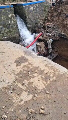 The scene shows water cascading from a stone structure into a hole. A red pipe lies in the path of the water flow. There are pieces of concrete scattered on the ground, along with some small rocks. The surrounding area appears to be muddy with a deep ravine on the right side. Some people can be seen standing on the structure.