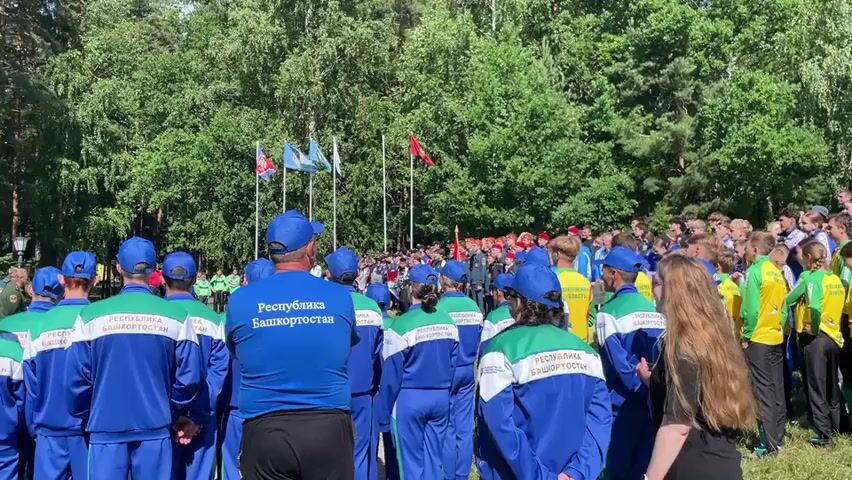 The video shows an outdoor gathering of people, likely a sporting event or a youth camp. A large group of young people, mostly teenagers, are gathered in a semi-circle. Many are wearing matching tracksuits in blue, green, and white, some with the inscription "REPUBLIKA BASHKORTOSTAN" on the back, indicating they are from the Republic of Bashkortostan. Others are wearing blue tracksuits with "RUSSIA" written on them. There's a stage set up on the left side of the frame with a banner that reads "VOSPITANIE DETEY" (Education of Children). A few adults appear to be speaking on the stage. In the background, there are several flags on poles, including what looks like the Russian flag and regional flags. The environment is a park or forest area with mature trees and green grass. The overall atmosphere seems formal, possibly an opening ceremony or a presentation.