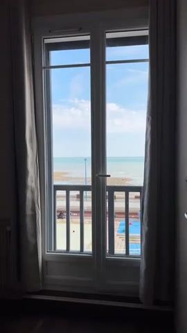 A full shot shows a double glass door with a white frame, flanked by beige curtains. The door opens to a view of the ocean under a blue sky with scattered clouds. In the middle ground, there's a railing, and beyond it, a beach with some scattered objects. The floor inside the room is dark wood.