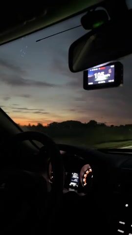 The video shows the interior of a car driving at dusk. The dashboard displays the speed at 105 km/h. A dashcam mounted on the windshield shows the same speed. The sky is visible through the windshield with clouds and the sun setting on the horizon. The road appears to be a highway or a main road with trees and buildings in the distance. The lighting suggests it is either early morning or late evening.