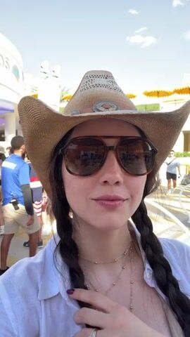 A woman is taking a selfie at what appears to be a pool party. She is wearing a straw cowboy hat, large brown sunglasses, a white blouse and gold necklaces. She has long, dark hair in braids. Behind her, there are many people milling around a pool. There are orange umbrellas providing shade and a structure with the word "Flamingo" on it. The sky is blue and sunny.
