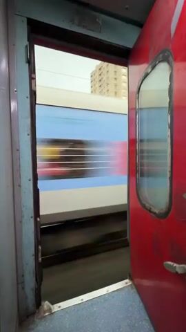 The video opens from inside a train compartment, looking out of an open doorway. The door is red and has a window with a rubber seal. The inside of the train compartment appears to have blue flooring and light-colored walls. Outside the doorway, another train is passing by. The passing train is blue and white with horizontal lines. The video then transitions to show the view from the open doorway as the train moves. The landscape outside is blurred due to the motion. Green trees and vegetation are visible, along with some buildings and structures. The sky is overcast. The camera is positioned at a slight downward angle, showing the train tracks. The video is short and appears to be a first-person perspective from someone standing in the doorway of a moving train.