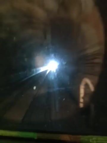 The video displays a dark, reflective surface with a bright light source in the center. The surface appears to be scratched or cracked, with streaks of light emanating from the bright spot. There are a few small, light-colored particles scattered on the surface. In the upper portion of the frame, a blurred, circular reflection is visible, suggesting a lens or an eye. The lower portion of the frame shows a greenish-colored edge.