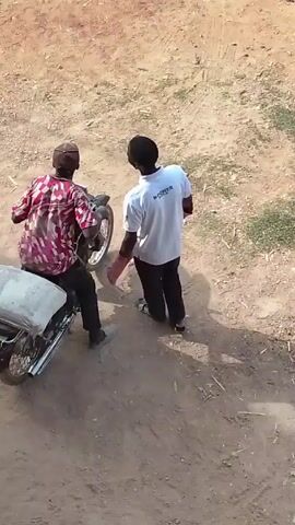 Two individuals are observed in an outdoor setting with a dirt ground surface. One individual is seated on a parked motorcycle, dressed in a red and pink patterned shirt, dark trousers, and a baseball cap. A white bag or covering is on the motorcycle's passenger seat. The second individual, wearing a white t-shirt with the word "N-POWER" visible, dark trousers, and sandals, is standing and walking away from the motorcycle. They have what appears to be a red glove or covering on one hand. The scene is captured from an elevated viewpoint, providing a clear view of the immediate surroundings and the interaction between the two individuals. The motorcycle's exhaust can be seen emitting a small amount of smoke.