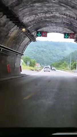 The video begins with a view from inside a vehicle looking forward through a tunnel. On the tunnel ceiling, overhead signs display green arrows pointing downwards, indicating traffic flow, and a circular sign with the number 80 inside, signifying a speed limit of 80. As the vehicle exits the tunnel, the scene opens to a multi-lane highway. The sky is overcast with heavy clouds. Lush green, tree-covered mountains dominate the background, stretching as far as the eye can see. Several vehicles, including white vans and cars, are traveling in the same direction on the highway. A speed limit sign showing 90 is visible on the side of the road. The highway has clear lane markings, with solid and dashed white lines. A guardrail runs along the side of the road. The overall environment appears to be a rural or mountainous region.