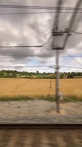 The video shows a view from a moving train. The train tracks are visible in the foreground, blurred due to motion. Beyond the tracks is a rural landscape with fields of golden crops, possibly wheat or barley. Trees and bushes dot the landscape, and in the distance, there are rolling hills and a cloudy sky. A power line pole with wires is visible, and there are a few other utility poles scattered in the distance. The motion blur indicates the perspective of someone traveling by train.