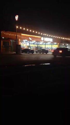 The scene is at night and depicts a Vostok store illuminated by string lights. Several cars are parked in front, and a small truck is further down. One car drives past the camera toward the right. The ground in front of the camera is wet, possibly from rain.