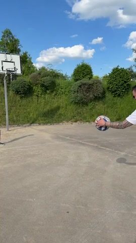 A man is seen shooting a basketball on an outdoor court on a sunny day. The man is wearing a white shirt, black shorts, and black shoes. He has tattoos on his arm and leg. The basketball has red, white, and blue markings.  The basketball hoop is white and black. The court is concrete and surrounded by grass and bushes. The sky is blue with white clouds. The man misses the shot.