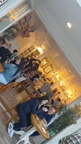 The video begins inside of a cafe. Several people are seated at tables. A woman is resting her head on the shoulder of a man seated next to her. The camera exits the cafe. Outside, a few people are walking past the cafe. Further out, there are more outdoor tables and umbrellas. Trees are visible as the camera pans upward.  The day appears to be overcast.