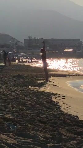 At 00:00, a person on a sandy beach prepares to do a flip with the ocean and a town in the background. At 00:01, the person completes the flip and lands in a crouched position. At 00:02, the person is in a push-up position. At 00:03, the person is still in the push-up position. At 00:04, the person starts to stand up.