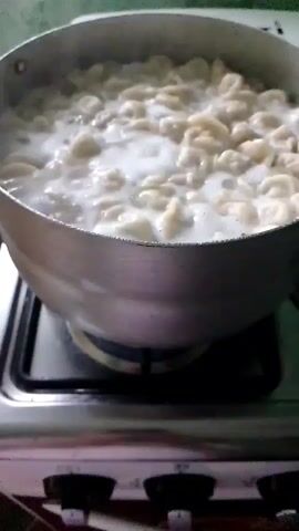 A pot of pasta is boiling on a stovetop. The pasta is a pale yellow color and is curved in shape, resembling macaroni. The water is bubbling vigorously, with some foam on the surface. The pot is made of silver-colored metal and is sitting on a black gas stove. The stove has three control knobs at the front, which are black. A white surface is visible behind the pot, possibly a backsplash or the top of the stove. The overall environment appears to be a kitchen.