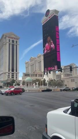 The video captures a street view in Las Vegas, Nevada, focused on the iconic Caesars Palace. From a slightly low angle, taken from inside a vehicle, the scene presents Caesars Palace with its distinct architecture on the left, and the famous digital billboard displaying "The A’ja Wilson Show" on the right. The billboard features a rotating set of images of basketball player A’ja Wilson. The background consists of a clear, blue sky interspersed with scattered clouds. 

Traffic flows along the intersection in front of the camera, which includes various vehicles such as a red car, a black car, a silver SUV, and a black SUV. To the right, the front part of a white van is visible, offering a glimpse of the side mirror and front panel. The lighting suggests a bright, sunny day.