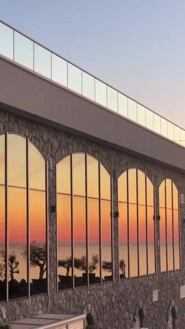 The video depicts a scenic view of a sunset over the sea. Initially, the camera focuses on a building with large, arched windows reflecting the colorful sunset, composed of hues of orange, pink, and yellow. The building's lower section is constructed of stone, and it features a glass-paneled balcony at the top. As the video progresses, the camera pans towards the right, revealing the vast expanse of the sea and the vivid sunset sky. The camera then stops to show street lights, trees, a swingset, and a sign which reads "Ресторан Царевић," which may be the name of a restaurant in the area. A few people are visible on the swingset. A car can be seen parked in the background as well.