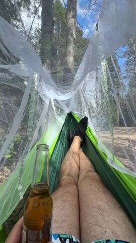 A person lies in a green hammock, with black socks on their feet, holding a bottle of amber liquid, possibly beer. A white mosquito net surrounds the hammock. The background features trees and a blue sky with light clouds. The perspective is from inside the hammock, looking out and up.