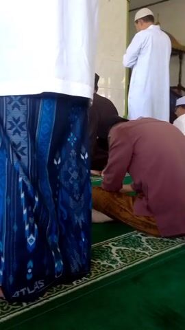 The video shows a group of men praying inside a mosque. They are dressed in traditional Islamic clothing, including long robes and skullcaps. The mosque has a green carpet with a decorative border, and the walls are white. Some of the men are standing, while others are kneeling or bowing in prayer. There is a wooden pulpit in the background. The lighting is natural and appears to be coming from windows. The video is filmed from a low angle, looking up at the praying men.