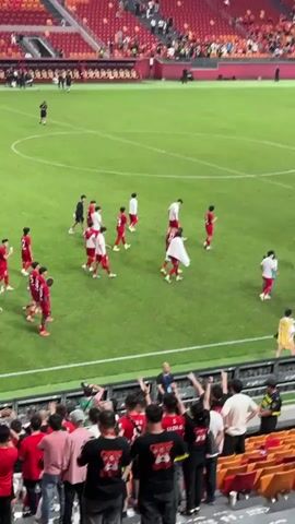 The video shows a soccer stadium with many fans in red and black clothing filling the stands. Players wearing red and white uniforms walk onto the green soccer field. The crowd cheers and waves. There are banners with writing on them in the stands.