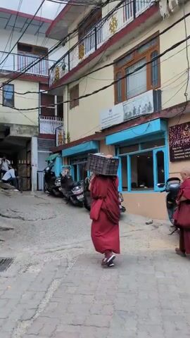 Two monks in maroon robes walk down a narrow, paved street lined with buildings. The monk on the left carries a dark crate on their head. The monk on the right carries a light-colored bag. Scooters are parked along the side of the street. Electrical wires are strung overhead. The buildings have balconies and windows. The sky is overcast.