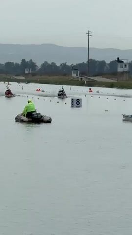 A wide shot captures a canoe race on a misty, overcast day. Several competitors are paddling in canoes, each marked with a number in a white square floating on the water. A person in a yellow vest operates a small inflatable boat in the foreground, acting as a course marshal or safety boat. The race appears to be taking place on a calm body of water with a distant shoreline lined with trees and structures. The overall atmosphere is serene and focused on the sporting event.