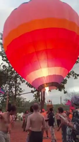 The video shows a partially inflated hot air balloon on the ground with people milling around it. The balloon is brightly colored in a striped pattern with white, yellow, red, pink, purple, and blue. There is a small fire coming from the center of the balloon. Most of the people are shirtless and seem to be watching the balloon. There are trees around the edge of the scene. The sky is overcast. The video is shaky.