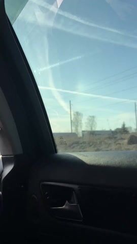The video is shot from inside a moving car, showing the view through the passenger side window. The sky is blue with contrails from airplanes. A speed limit sign with "70" is visible along with utility poles and wires. An arched building or structure can be seen in the distance. The interior of the car is dark, and the door handle is visible.