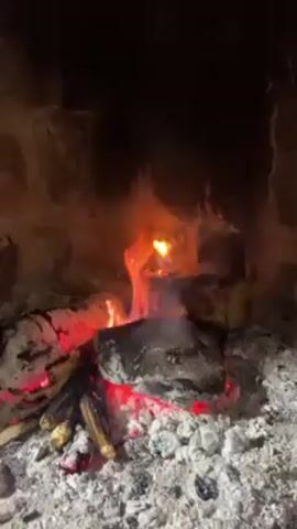 A fireplace filled with logs ablaze is the central element of this close-up shot. Flames dance upwards, illuminating the interior of the fireplace. Several ears of corn rest at the base of the burning logs, nestled among the embers and ash. The fireplace appears to be made of stone, with soot stains visible on the interior walls. There is no one visible in the video, only the crackling fire and the roasting corn.