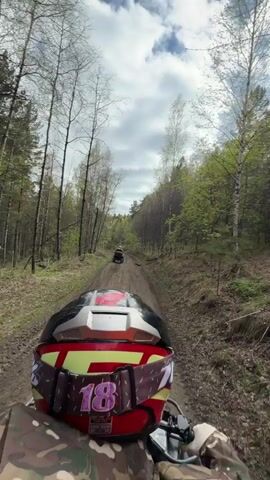 A first-person perspective video shows someone riding an ATV on a dirt road through a forest. The rider is wearing a camo jacket and a helmet with red, yellow, and white coloring, and the number 18 is visible on the goggle strap. The road is narrow, with trees on both sides. In the distance, another ATV is moving along the same path. The sky is overcast.