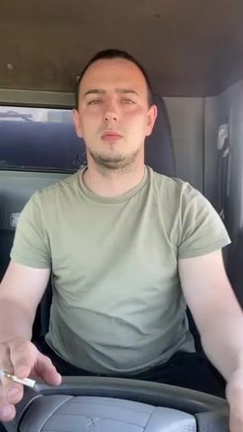 A man in a light green t-shirt is sitting in the driver's seat of a vehicle, possibly a truck. He has short brown hair and a stubble beard. He is smoking a cigarette. He looks towards the camera and then away, exhaling smoke. A wedding ring is visible on his left hand. The interior of the vehicle is visible, including the steering wheel, window, and dashboard. Background music is playing.