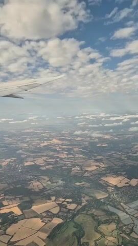 The video shows an aerial view from an airplane window. The wing of the plane is visible in the upper left corner. Below, a patchwork of agricultural fields, some golden and some green, stretches across the landscape. Scattered throughout the fields are small clusters of buildings, suggesting villages or towns. A winding river or body of water is visible, with some fields appearing to be flooded or recently irrigated. The sky is blue with scattered white clouds. The overall impression is of a rural landscape seen from a high altitude.