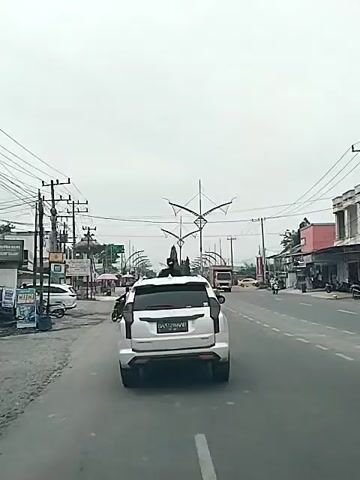 A white Mitsubishi Pajero Sport SUV is driving down a paved road, as seen from behind. The road is flanked by electrical poles, shops, and businesses. There are decorative streetlights resembling abstract birds along the road. Other vehicles, including motorcycles and a truck, are visible on the road. The sky is overcast. A piece of trash blows across the road. The video appears to be taken from inside another vehicle following the SUV.