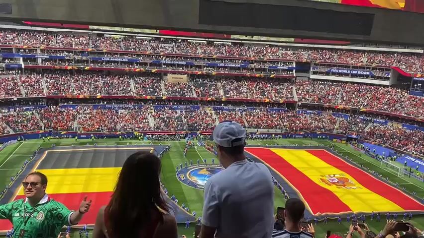 A high-angle, wide shot shows a packed soccer stadium. Two large flags, one predominantly black and yellow and the other red and yellow with an emblem, are laid out on the green field. Spectators fill the tiered seating all around the field. In the foreground, from the perspective of a spectator, are the backs of two people, a man and a woman, looking out at the field. A man wearing a green soccer jersey and sunglasses, with his thumb up, is visible on the left side of the frame.