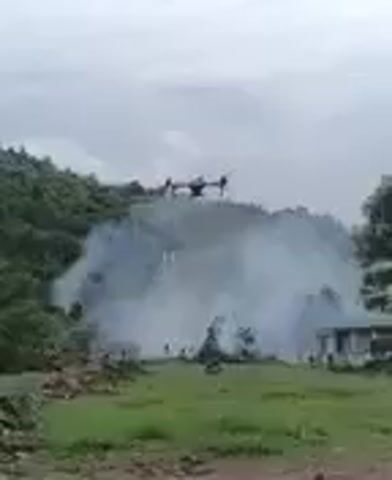 A drone hovers in the air, its rotors spinning as it emits a red light from its underside. Below the drone, a plume of white smoke billows upwards, partially obscuring the landscape. In the background, a hillside covered in green vegetation rises steeply. To the right of the smoke, a small, white building with a dark roof is visible. In the foreground, the ground is covered in green grass, with some debris and rocks scattered about. Several figures, appearing to be people, are partially visible standing amidst the smoke and vegetation. The sky is overcast with a muted, grayish hue.