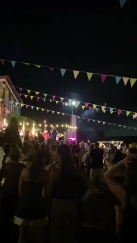 A large crowd of people are gathered outdoors at night for what appears to be an event or festival. String lights and colorful triangular pennant banners are strung across the scene, creating a festive atmosphere. In the background, a stage is set up with performers, and lights are illuminating the area. The crowd is mostly facing the stage, and many people are dancing or socializing. The overall environment suggests a lively outdoor celebration at night.