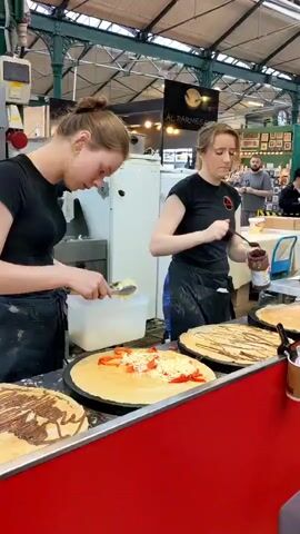 Two young women in black t-shirts and black aprons are preparing crepes at a food stall. The woman on the left is using tongs to add shredded cheese and chopped red bell peppers to a crepe on a round griddle. She is holding a white plastic container in her left hand. The woman on the right is spreading chocolate spread onto a crepe with a spatula, using a jar of chocolate spread. Both women appear to be focused on their tasks. In the background, a man in a black t-shirt is visible, holding a bag. There are several crepes on display in the foreground, some with chocolate drizzle. The environment appears to be an indoor market with a high ceiling and exposed beams. A sign above the stall reads "AL PARMESANO."