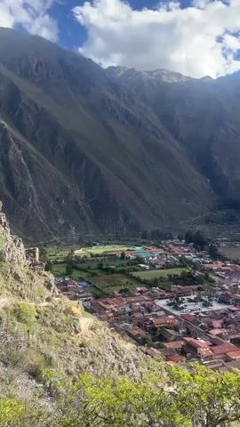 The video shows a town nestled in a valley, surrounded by large mountains under a partly cloudy sky. The town has buildings with red-tiled roofs, and there's a football field visible. On one side of the valley, there are terraced structures built into the mountainside. The sun shines through the clouds, casting rays of light onto the landscape. The video is taken from an elevated position, looking down at the town. A bush or small tree is visible in the foreground.