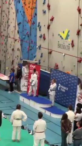 The video shows a woman in a judo gi, a white uniform with a blue belt, standing on the first-place tier of a podium. Another woman, also in a judo gi with a blue belt, stands beside the podium clapping. A climbing wall is visible in the background. A banner for France Judo stands next to the podium. Other people are standing around the edge of the room. The woman on the first-place tier bows her head.