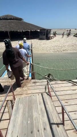 This video is shot from a wooden pier. In the foreground, a person is walking away from the camera, up a set of wooden stairs. There are other people walking ahead of them, also on the pier. The pier leads to a sandy beach with several buildings, including a large thatched-roof structure and some smaller white structures. The water next to the pier is clear and appears to be shallow. The sky is clear and blue. There are people walking on the beach in the background.