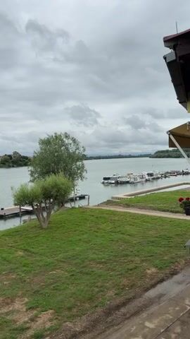 The video pans across a cloudy sky over a wide, calm river. On the river, several boats are moored to a dock. A small, sculpted tree stands on a grassy bank in the foreground. The riverbank is lined with more trees and buildings in the distance.