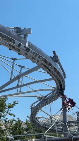 A vertical video captures a roller coaster ride against a clear blue sky. The ride features a grey and black track that curves and loops. A red roller coaster car with two passengers is visible, suspended from the track. The passengers appear to be seated side-by-side. The car is in motion, with the track extending above and below it. In the background, trees and mountainous terrain can be seen. The footage includes a close-up of the car and its occupants, followed by a wider shot of the roller coaster and the surrounding landscape.