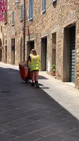 This video captures a person with long blonde hair walking away from the camera down a narrow cobblestone street. They are wearing a bright yellow safety vest over a dark top and a dark skirt or shorts. They are pushing an orange wheeled cart filled with items. The street is lined with rustic stone buildings featuring arched doorways, shuttered windows, and small potted plants. A flag with red and white stripes is visible on a building to the left. The sunlight casts distinct shadows on the pavement, suggesting it is a bright, sunny day, likely in the morning or afternoon. The setting appears to be a historic European town, consistent with the provided location context of San Quirico d'Orcia, Italy. The overall mood is one of daily activity in a charming, ancient urban environment. There is no visible text in the video.