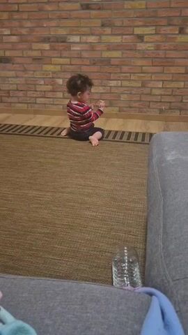 A young child, wearing a red and white striped shirt and dark shorts, is sitting on a brown rug. The child is facing a brick wall.  A gray couch with a textured surface is visible in the foreground, and there is a glass of water sitting on the couch. The child initially looks down, fiddling with something in their hands, then looks up at the camera and smiles brightly, holding their hands up. The room appears to be an indoor living space with a modern design. The child is barefoot.  The lighting is warm and seems to be artificial. There are hardwood floor accents near the base of the brick wall.  Music is playing in the background.