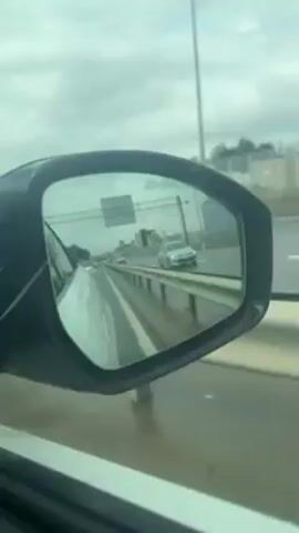 The video shows the view from a car side mirror on a rainy day. The mirror reflects a highway with other cars and buildings in the distance. The sky is overcast, and the road is wet. The camera appears to be moving, suggesting the car is in motion.