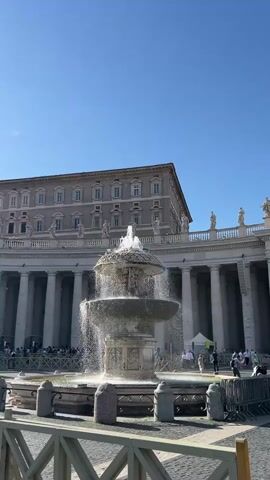 The video captures a sunny day at St. Peter's Square in Vatican City. The first shot shows a large, ornate fountain spraying water, with a grand, multi-story building and a colonnade visible in the background under a clear blue sky. The second shot pans across the square, revealing St. Peter's Basilica with its iconic dome, more of the colonnade, and a large crowd of people walking around. The sun is bright in the sky, and there are a few scattered clouds. Some temporary barriers are set up in the square.