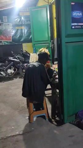 The video shows a person sitting on a stool in front of a food stall at night. The person is wearing a black jacket with "MUHAMMAD ALI" written on the back and a baseball cap. They are holding a bottle and appear to be eating or drinking. Behind the person, a motorcycle is parked, and a green food stall with a yellow frame is visible. A banner behind the stall advertises "Rujak & Gado" with images of food. The setting appears to be an outdoor market or street food area in Satriamekar, Indonesia, illuminated by artificial lights. There is a sign for "DRAVA" on the right side of the frame. The overall mood is casual and suggests a late-night meal or snack.